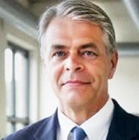 Professional headshot of a middle-aged man with gray hair wearing a dark suit and blue tie